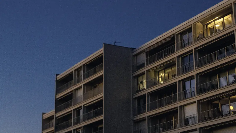 A mid-rise apartment building with balconies at dusk, a few windows lit.